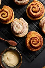 partially frosted batch cinnamon rolls spread out on a countter alongside  a bowl of frosting